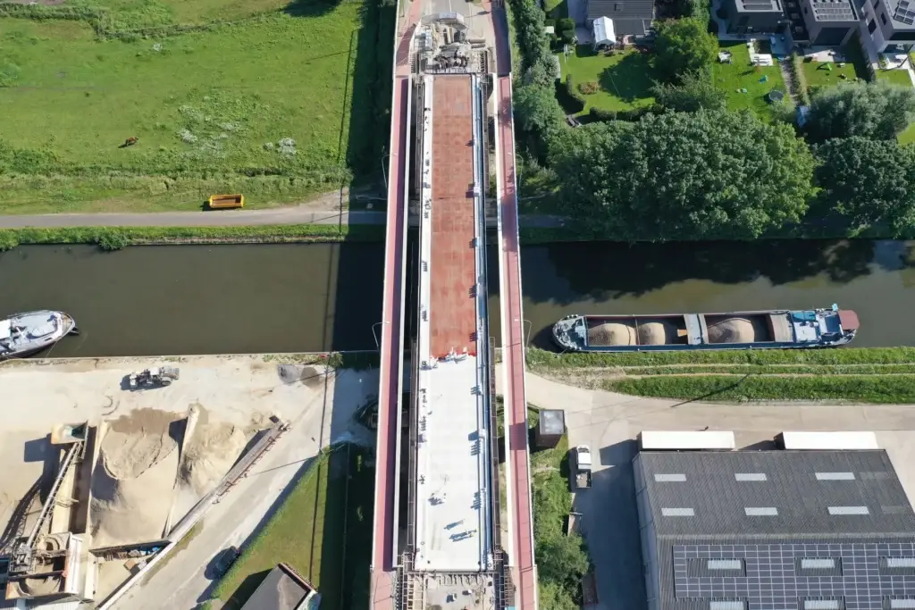 Luchtfoto van een brug in renovatie boven een kanaal, met werfmaterialen en arbeidzones op het brugdek. Aan beide zijden van het water liggen schepen aangemeerd, omringd door groen en nabijgelegen bedrijfsgebouwen.
