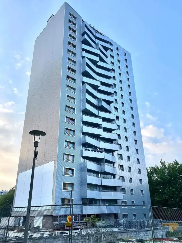 Hoog residentieel gebouw met vernieuwde gevel en opvallende zigzag‑vormige balkons, gefotografeerd vanop straatniveau. Het gebouw heeft een moderne, zilverkleurige afwerking en staat naast een omheinde werfzone met materiaal en hekwerk.
