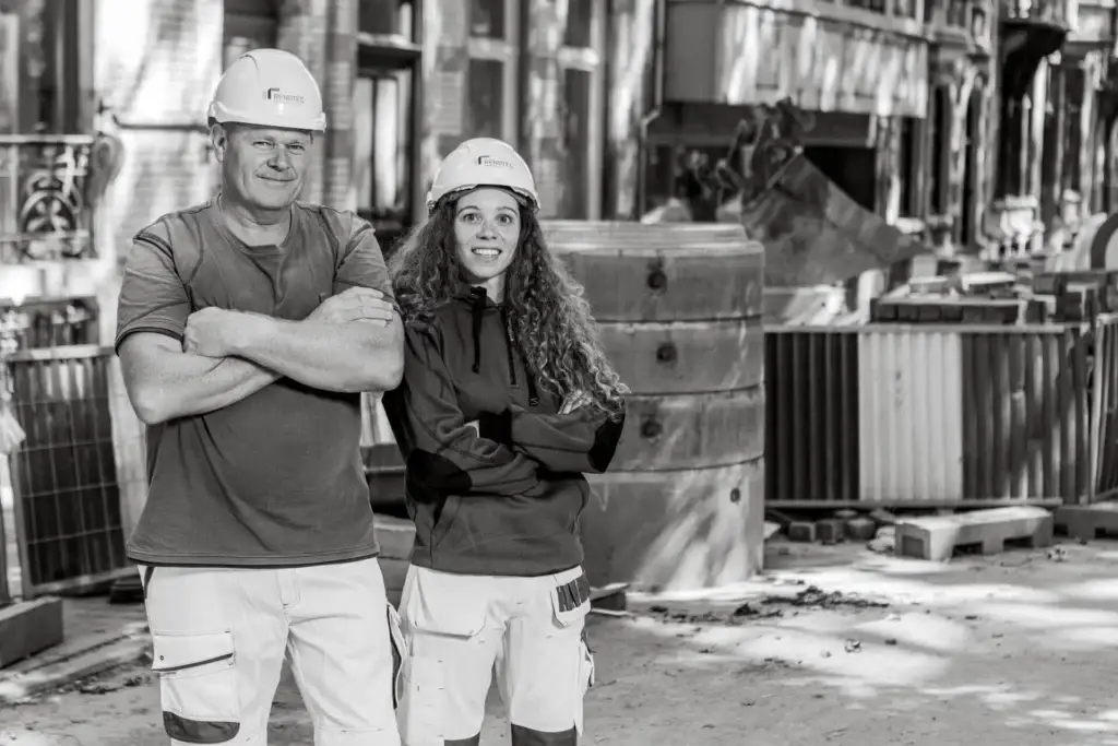 Twee personen in werkkledij en veiligheidshelm staan met gekruiste armen op een stedelijke werfzone tussen bouwmaterialen, tijdelijke afschermingen en historische gevels, gebruikt als wervings‑ en projectvisual binnen een renovatie‑ en restauratiecontext.