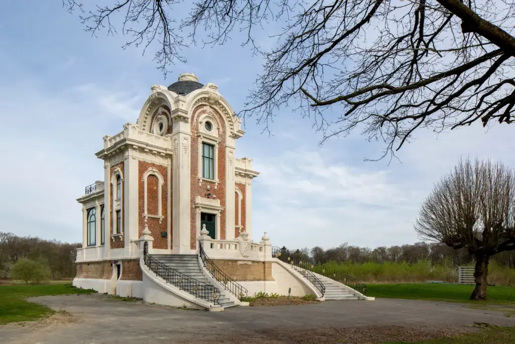 Historisch gebouw met monumentale trap en rijk uitgewerkte gevel, omringd door een open groen landschap en bomen. Symmetrische architectuur met decoratieve elementen en grote ramen.
