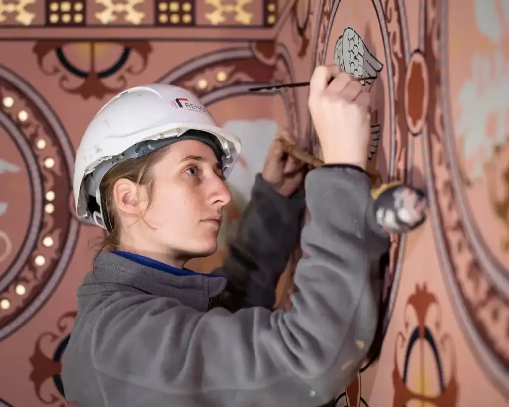 Restauratiemedewerker die met een fijn penseel decoratieve muurschilderingen bijwerkt op een historisch interieur, met aandacht voor lijnwerk en ornamentale details.