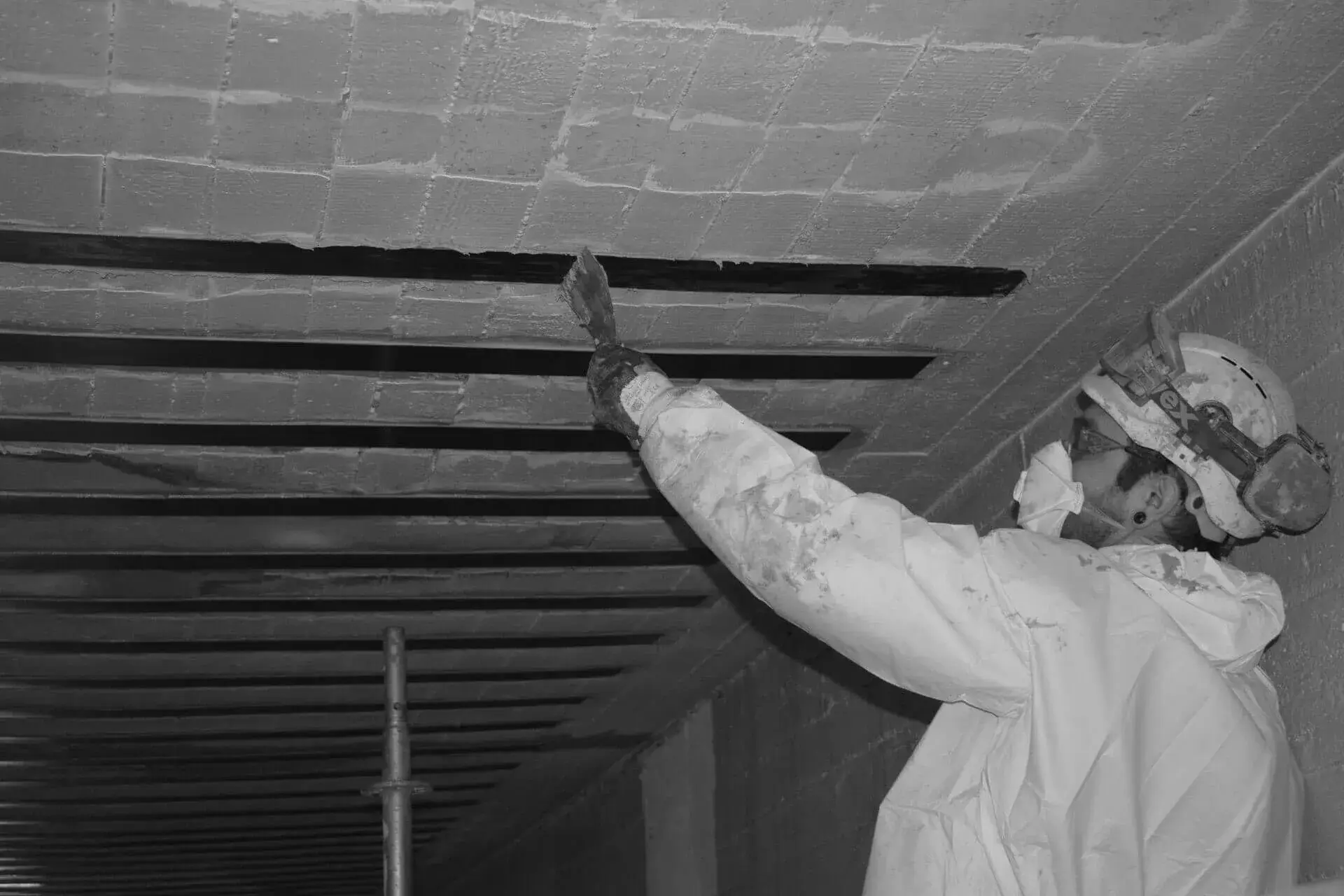 Medewerker in beschermende overall brengt gelijmde wapening aan op een betonnen plafond, waarbij meerdere sleuven voorbereid en gevuld worden tijdens structurele versterkings‑ en renovatiewerken.