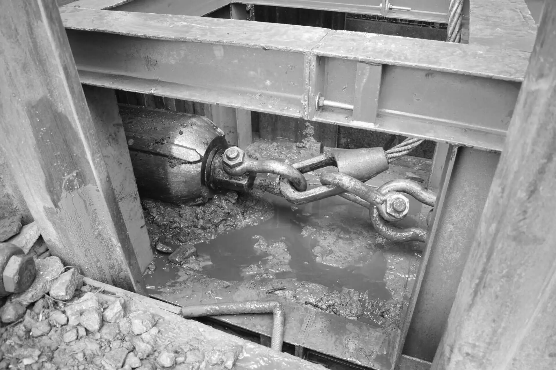 Pipe‑bursting installatie in een beschoeide sleuf, met een trekconus en zware kettingverbinding die door de modderige werkzone loopt tijdens de vervanging van een ondergrondse leiding.