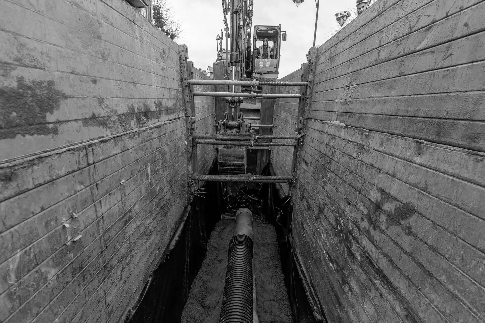 Open sleuf met beschoeiing waarin een graafmachine een grote riooleiding positioneert en de bedding aanvult, onderdeel van rioolrenovatiewerken en ondergrondse infrastructuurwerken.